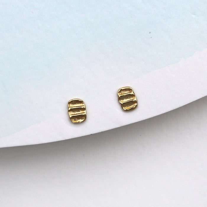 A pair of small gold stud earrings with a textured, ribbed design, displayed on a white card against a light blue background.