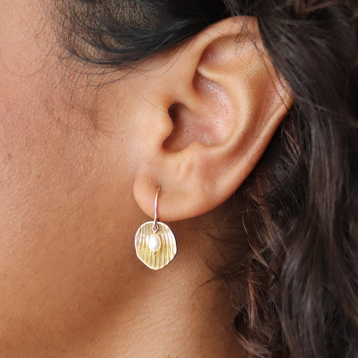 A close-up of a person's ear wearing a small gold hoop earring with a textured, shell-like gold dangle and a tiny white pearl accent.