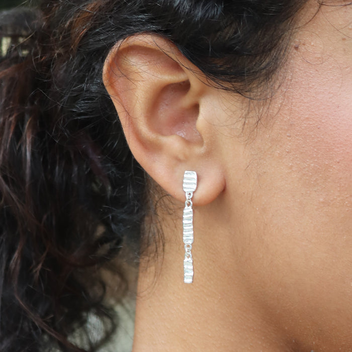A close-up of a person's ear wearing a delicate silver drop earring with textured rectangle links.
