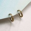 A pair of small textured gold hoop earrings resting on a curved, light blue paper edge against a textured off-white background.